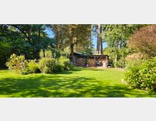 Großer Garten mit grünem Rasen, umgeben von dichtem Baumbestand und Sträuchern. Im Hintergrund ein Schuppen mit Holzlager und Gartenutensilien, daneben ein kleiner Grillbereich.