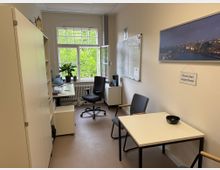 Kleines Büro mit Schreibtisch, Bürostuhl, Regal und zwei Besucherstühlen an einem kleinen Tisch. Ein großes Fenster mit Blick auf grüne Bäume lässt viel Tageslicht herein, während eine weiße Pinnwand und ein Stadtbild an der Wand hängen.