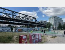 Eine mit Graffiti verzierte stählerne Eisenbahnbrücke mit umliegenden Betonwänden, die ebenfalls bemalt sind. Im Vordergrund befindet sich eine asphaltierte Fläche mit einem kleinen Parkbereich, einer rot markierten Wasserentnahmestelle und Fußgängern; rechts sind moderne Wohngebäude und Bäume sichtbar.