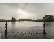 Holzsteg mit dekorativen Geländerpfosten an einem ruhigen See bei Regen; im Hintergrund sind Bäume und dichte Vegetation unter bewölktem Himmel sichtbar.