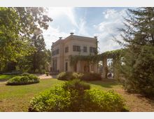 Kleines helles Gebäude mit klassizistischen Architekturelementen, wie symmetrischen Fenstern und einer überdachten Terrasse, die mit Kletterpflanzen bewachsen ist. Umgeben von einem gepflegten Garten mit Rasen, Büschen und hohen Bäumen im Hintergrund, unter klarem Himmel.