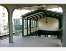 Überdachte Treppenabgangskonstruktion an einer erhöhten U-Bahnstation mit grünem Metallrahmen und großen Glasfenstern; im Vordergrund ein offener Bereich mit einer Metalltür und Blick auf umliegende Wohnhäuser.
