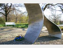Metallskulptur in einem Park mit geschwungenen Formen und eingraviertem Text, umgeben von Bäumen und Pflanzen. Im Vordergrund liegt ein Blumenkranz, und im Hintergrund steht eine weiße Parkbank auf einem gepflasterten Weg.