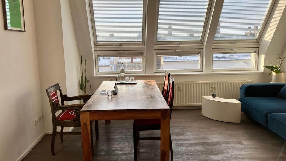 Bright attic-style living and dining area with slanted ceiling windows fitted with blinds, overlooking rooftops. A wooden dining table with chairs sits in the center, with a blue sofa and curved white side table beside a radiator and potted plants.