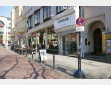 Straßenszene mit mehreren Geschäften, darunter ein Juwelier und ein Café namens „Balzac Coffee“. Links stehen Außenbestuhlung mit Tischen und Stühlen, teilweise besetzt, daneben eine gepflasterte Fußgängerzone. Die Gebäude zeigen eine Mischung aus historischer und moderner Architektur.