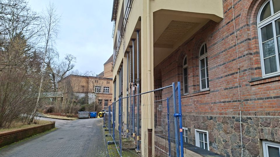 Schmaler gepflasterter Weg entlang eines Backsteingebäudes mit Rundbogenfenstern und Natursteinsockel; davor steht ein Baugerüst mit blauem Bauzaun unter einem auskragenden Vordach. Links säumen kahle Bäume und Sträucher den Weg, im Hintergrund sind weitere Backsteinbauten und Container zu sehen.