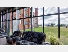 Innenansicht eines modernen Gebäudes mit großer Glasfassade, durch die ein Ziegelgebäude und eine Grünfläche sichtbar sind. Im Vordergrund stehen zwei schwarz-gemusterte Sessel und eine Pflanze, seitlich führt eine Metalltreppe nach oben.