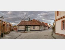 Kopfsteinpflasterstraße in einer kleinen Ortschaft mit historischen Häusern, darunter ein einstöckiges Gebäude mit rotem Ziegeldach und grüner Tür im Vordergrund. Links eine Backsteinmauer mit Straßenlaterne und rechts weitere Häuser, im Hintergrund der Kirchturm einer Kirche.