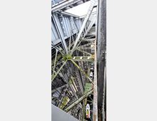 Metallische Konstruktion im Inneren eines Schiffshebewerks mit komplexen Stahlträgern und sichtbaren Seilzügen. Unten im Hebewerk sind kleine Boote zu erkennen, die auf Wasser schwimmen.