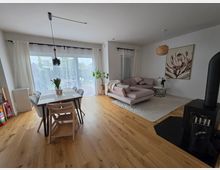 Offener Wohn- und Essbereich mit hellem Holzfußboden, Esstisch mit Stühlen vor großen Schiebetüren und weißen Vorhängen. Rechts eine L‑förmige Sofaecke auf Teppich mit Beistelltisch, Wandbild und Hänge-/Deckenleuchten sowie ein schwarzer Kaminofen.