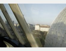 Blick durch schräge Glasfenster auf eine Stadtszene mit rechteckigen Wohngebäuden und einem kleinen Park im Vordergrund; rechts ein Teil einer modernen, kuppelförmigen Metallstruktur.