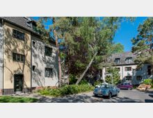 Mehrfamilienhäuser im Stil der Nachkriegsarchitektur aus verputztem Mauerwerk, umgeben von Bäumen und Grünflächen. Im Vordergrund eine ruhige Straße mit parkenden Autos und einem Straßenschild.