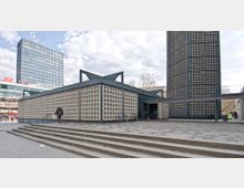 Eine moderne Kirche mit geometrischem Design und Wabenstrukturwänden, flankiert von einem hohen, fensterlosen Turm. Im Hintergrund ist ein Hochhaus mit dem Mercedes-Benz-Logo zu sehen, umgeben von einem urbanen Platz mit Treppen und Fußgängern.
