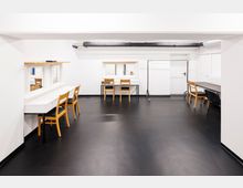 Ein heller Umkleideraum mit weißen Wänden, schwarzem Boden und mehreren Schminktischen mit Spiegeln. Holzelemente wie Stühle und Regale prägen den Raum, zusammen mit offenen Rohrleitungen an der Decke.