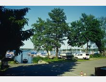 Ein Seeufer mit einer kleinen Anlegestelle, an der Segelboote vertäut sind. Im Vordergrund Bäume, ein geparkter Transporter und ein Boot auf einem Anhänger, dahinter Wasser und klare Sicht auf das andere Ufer.