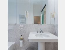 Modernes Badezimmer mit einem weißen Waschbecken auf einem Standfuß, einer Wand mit grauen Fliesen, einem großen Spiegel und einem Handtuchhalter. Im Hintergrund ist eine Tür und ein WC zu sehen.