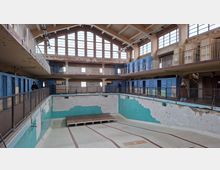 Ehemaliges Hallenbad mit leeren Becken und verblasster, türkisfarbener Fliesenverkleidung, umgeben von zwei Galerien mit blauen Umkleidekabinen und großen Fenstern, die im Obergeschoss viel Licht hereinlassen. Die Architektur weist sichtbare Alterungsspuren und abgenutzte Oberflächen auf.