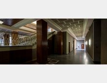 Innenansicht eines Gebäudes im Stil der 1950er oder 1960er Jahre mit dunklen Holzverkleidungen an den Säulen, einem Treppenaufgang mit verzierten Geländern aus goldfarbenem Metall und einem langen Flur mit hellen Wandlampen. Die Decke ist mit quadratischen Mustern dekoriert und der Boden mit dunklen Fliesen ausgelegt.