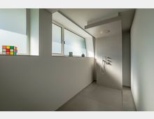 Minimalistisch gestaltetes Badezimmer mit einer bodengleichen Dusche auf der rechten Seite und großen, milchglasverkleideten Fenstern, die weiches Licht hereinlassen. Auf der Fensterbank steht ein Rubik's Cube neben kleinen Dekorobjekten.