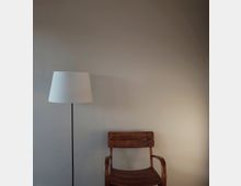 Eine minimalistische Innenraumaufnahme mit einer einfachen Stehlampe mit weißem Lampenschirm links und einem hölzernen Stuhl mit Armlehnen rechts vor einer beigen Wand.
