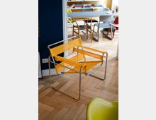 Ein modernes Interieur mit einem gelben Designerstuhl aus transparentem Material und einem verchromten Metallrahmen, auf einem Parkettboden. Im Hintergrund ein Esstisch mit verschiedenen Stühlen und ein Bücherregal an einer gelb gestrichenen Wand.