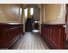 Ein historisches Treppenhaus mit verzierten Stuckdetails und dunkelroten Holzvertäfelungen an den Wänden. Der Boden ist mit einem gemusterten Fliesenmosaik gestaltet, und im Hintergrund führt eine Treppe zu einer oberen Ebene, während eine hölzerne Tür mit Fenster am Ende des Gangs sichtbar ist.