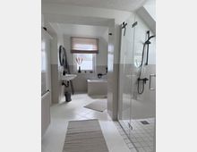 Helles Badezimmer mit weiß gefliesten Wänden und Böden, ausgestattet mit einer Glasduschkabine, einer Badewanne, einem Waschbecken mit rundem Spiegel und einem Fenster mit halbtransparentem Rollo. Schwarze Armaturen und beige Teppichläufer ergänzen das minimalistische Design.