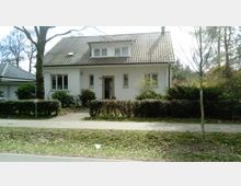 Einfamilienhaus mit weißer Fassade und Satteldach, umgeben von Sträuchern und Bäumen. Vor dem Haus führt ein gepflasterter Weg zu einer Eingangstür, daneben erstreckt sich Rasen mit einer Hecke zur Straße hin.