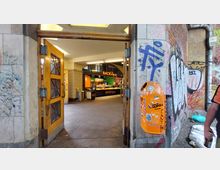 Eingang zu einem Gebäude mit doppelflügeliger Holztür, dahinter ein kleiner Verkaufsbereich einer Bäckerei mit der Aufschrift „BACKBaron“. Rechts außen ist eine orange Mülltonne mit Graffiti und daneben eine mit Graffiti besprühte Wand zu sehen.