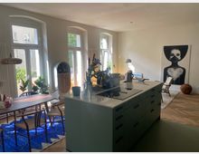 Moderner Wohnraum mit offener Küche, großem Esstisch und Stühlen auf blauem Teppich. Helle Fensterfronten mit Blick ins Grüne, minimalistisches Design, Wandkunst und Dekorationselemente verleihen dem Raum eine einladende Atmosphäre.