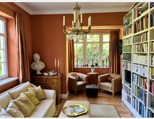 Gemütliches Wohnzimmer mit terrakottafarbenen Wänden, großem Kronleuchter und hohen Fenstern mit schweren Vorhängen. Im Raum stehen eine helle Couch, zwei Sessel mit Beistelltisch sowie eine lange, deckenhohe Bücherwand; Holzboden und ein Teppich ergänzen die Sitzecke.