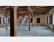 Offener Dachboden mit sichtbaren Holzbalken, Holzdielenboden und Backsteinwänden. Im Zentrum steht eine Holzleiter, umgeben von einer kunstvollen Installation aus gespannten Fäden.