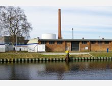 Backsteingebäude mit Flachdach und Schornstein an einem Kanal, umgeben von einer Wiese und einzelnen modernen Gebäuden im Hintergrund. Im Vordergrund befindet sich eine befestigte Uferkante mit Pollern.