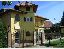 Gelbes Gebäude im mediterranen Stil mit roten Ziegeldächern, Rundbogenfenstern und grünen Fensterläden. Vor dem Haus befindet sich ein schmiedeeiserner Zaun mit Eingangstor, daneben ein Schild, das auf eine Pension hinweist.