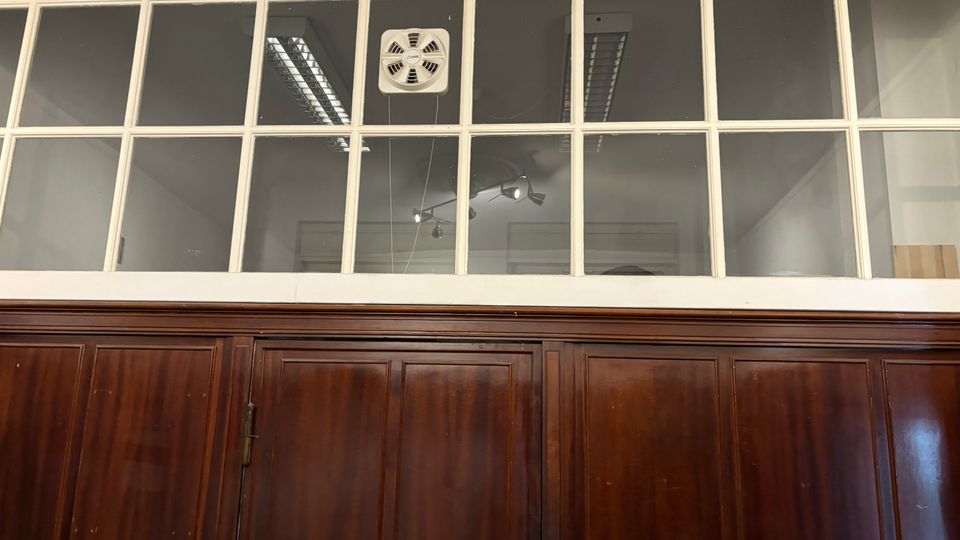 Innenansicht eines Raums mit dunkler Holzvertäfelung und einer darüberliegenden Glasfensterreihe in weißem Rahmen, die in einen angrenzenden Bereich blickt. Hinter den Scheiben sind Deckenleuchten und ein an der Decke montiertes Gerät sichtbar.