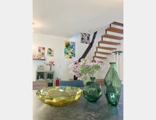 Ein moderner Wohnraum mit einer offenen Holztreppe an der Wand und abstrakten Kunstwerken an den Wänden. Im Vordergrund stehen eine gelbe Glasschale, zwei grüne Glasvasen und eine Vase mit lilafarbenen Blumen auf einem Tisch.