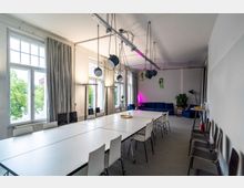 Heller Konferenz- und Arbeitsraum mit langen weißen Tischen und Stühlen, hohen Fenstern mit grauen Vorhängen und mehreren hängenden Deckenlampen. Am hinteren Ende stehen blaue Sofas neben Pflanzen, ergänzt durch eine pinke Akzentbeleuchtung.