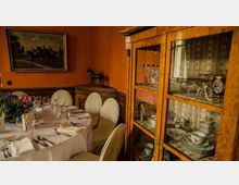 Ein gemütlicher Essbereich mit einer runden, weiß gedeckten Tafel, umgeben von gepolsterten Stühlen. Im Hintergrund stehen eine antike Vitrine mit Geschirr und Gläsern sowie ein Sideboard mit einer Pferdeskulptur, vor einer orangefarbenen Wand mit einem Gemälde.