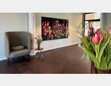 Wohnbereich mit dunklem Holzboden, einer hellgrauen Wand und moderner Dekoration. Im Vordergrund ein Blumenstrauß aus roten und rosa Tulpen; im Hintergrund ein Sessel, ein kleiner Beistelltisch mit Vase und ein großes Wandbild mit Tulpenmotiv.