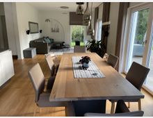 Essbereich und Wohnzimmer in einem modernen Haus mit hellem Holzboden, einem Esstisch aus Holz und gepolsterten Stühlen. Große Fenster lassen viel Tageslicht herein, im Hintergrund befindet sich eine graue Couch und eine Stehleuchte.