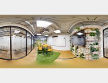 Moderner Besprechungsraum mit Glaswänden, großen Whiteboards und einer grünen Wand aus Holzpaletten mit Pflanzen. Eine Sitzgruppe mit gelben Stühlen steht auf einem Teppich in Grasoptik, die Decke zeigt sichtbare Lüftungsrohre. Ein Wandbild mit Naturmotiv ergänzt das Design.