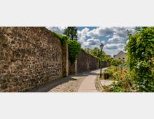 Eine gepflasterte Gehwegallee an einer alten Steinmauer, die mit Efeu bewachsen ist, flankiert von Blumenbeeten und einer Laterne. Im Hintergrund sind Bäume und Wohnhäuser unter einem teils wolkigen Himmel zu sehen.