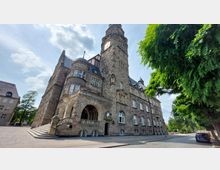 Historisches Gebäude aus Sandstein mit einem hohen Turm und einer Uhr, großen Rundbogenfenstern, verzierter Balustrade und geschwungenen Treppen. Rechts davon stehen grüne Bäume entlang einer gepflasterten Straße.