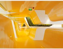 Futuristischer Innenraum mit geschwungenen gelben und weißen Wänden und Decken, durchsetzt mit Punktmustern. Der Raum verfügt über glatte Oberflächen, Glasabtrennungen und eine Wand mit grüner Bepflanzung im Hintergrund.