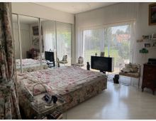 Schlafzimmer mit großem Doppelbett und floraler Bettwäsche, bodentiefen Fenstern mit weißen Vorhängen, die den Blick auf einen Garten ermöglichen. An der linken Wand ein großer Kleiderschrank mit Spiegeltüren, daneben ein kleiner Fernseher sowie diverse Möbelstücke und Dekorationen.