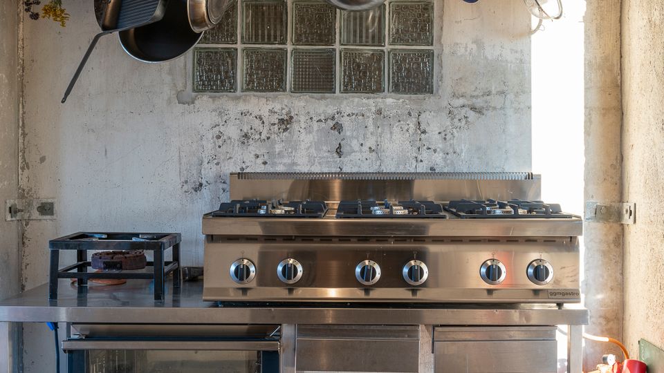 Industrieküche mit Edelstahlarbeitsfläche, einem großen Gasherd mit sechs Brennern und einem Ofen darunter. Oberhalb hängt an einem Metallgitter Kochgeschirr wie Töpfe, Pfannen und Kannen, während der Hintergrund aus Glasbausteinfeldern und einer Betonwand besteht.