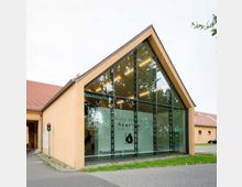 Modernes Gebäude in einem Satteldachstil mit großer Glasfassade, die den Blick auf eine innenliegende Ausstellung freigibt. Der angrenzende Bereich besteht aus gelb verputzten Wänden und einem roten Ziegeldach, das zu einem Innenhof führt; kleine Grünflächen und asphaltierte Wege befinden sich im Vordergrund.