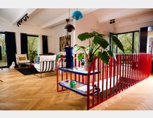 Helles, modernes Wohnzimmer mit Holzboden, roten Geländern um eine Treppe und großen Glasfenstern mit Blick auf eine grüne Außenlandschaft. Im Raum befinden sich Pflanzen, Designerstühle, ein Regal mit Dekorationsgegenständen und eine auffällige blaue Figur.