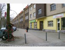 Straßenszene mit Reihenhäusern im historischen Stil, gekennzeichnet durch braune Fassaden mit gelben Fensterrahmen und roten Ziegeldächern. Im Vordergrund gelbe Gebäude mit einem Geschäft für Fußpflege und einer Fahrschule, daneben parkende Autos und ein Reifenständer an einem Baum.