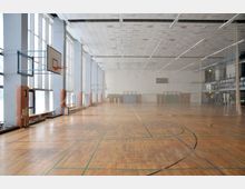 Eine große, helle Sporthalle mit Holzfußboden, markierten Spielfeldlinien und Basketballkörben an den Wänden. Große Fenster auf der linken Seite lassen viel Tageslicht herein, während im Hintergrund Pezzibälle, Matten und eine Handballtorwand sichtbar sind.